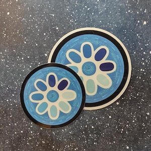 以下が含まれることがあります： 青い花の模様の丸いステッカーが2枚。花びらは、さまざまな青と白の色合いで、黒い縁取りのある水色の背景に配置されています。ステッカーは、暗い斑点のある表面にあります。