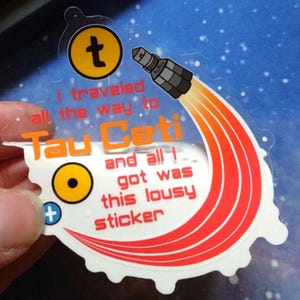 Puede incluir: Una pegatina transparente con un dise&ntilde;o de cohete y el texto "I traveled all the way to Tau Ceti and all I got was this lousy sticker." El dise&ntilde;o utiliza colores naranja y amarillo.
