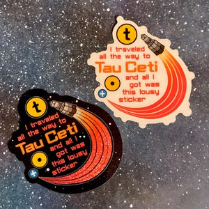 Puede incluir: Dos pegatinas con tem&aacute;tica espacial. Cada pegatina tiene el texto "I traveled all the way to Tau Ceti and all I got was this lousy sticker." Una pegatina es negra con purpurina, la otra es blanca.