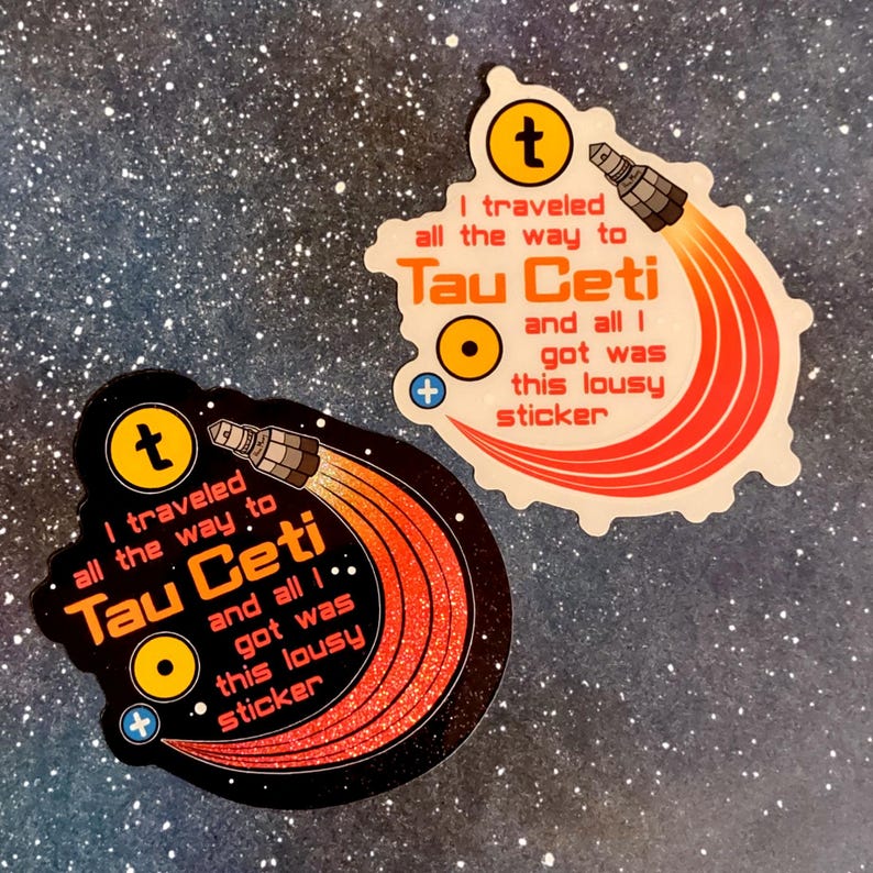 Puede incluir: Dos pegatinas con tem&aacute;tica espacial. Cada pegatina tiene el texto "I traveled all the way to Tau Ceti and all I got was this lousy sticker." Una pegatina es negra con purpurina, la otra es blanca.