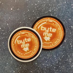 以下が含まれることがあります： オレンジと白のデザインの丸いステッカーが2つ。ステッカーには、白い文字で「byte me」とハートのシンボルが描かれています。黒と白のボーダーがあります。