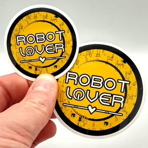 以下が含まれることがあります： 黄色い背景に白文字で「ROBOT LOVER」と書かれた2つの丸いステッカー。ステッカーは黒い縁取りと白い輪郭線があります。「LOVER」の下には小さなハートがあります。