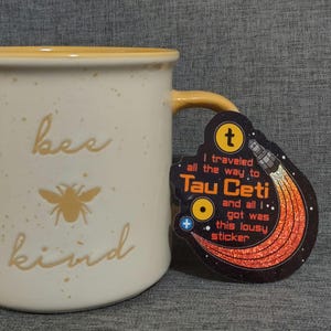 Puede incluir: Taza de cer&aacute;mica color crema con borde y asa amarillos. La taza presenta las palabras "bee kind" y un gr&aacute;fico de abeja. Una pegatina con el texto "I traveled all the way to Tau Ceti and all I got was this lousy sticker" est&aacute; adjunta.