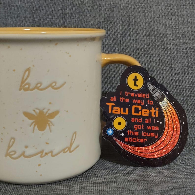 Puede incluir: Taza de cer&aacute;mica color crema con borde y asa amarillos. La taza presenta las palabras "bee kind" y un gr&aacute;fico de abeja. Una pegatina con el texto "I traveled all the way to Tau Ceti and all I got was this lousy sticker" est&aacute; adjunta.