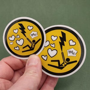 以下が含まれることがあります： 黄色い背景と黒い縁取りの丸いステッカー2枚。各ステッカーには、稲妻に打たれた人物が描かれ、白いハートで囲まれています。「VAH！」というテキストが吹き出しの中に表示されています。