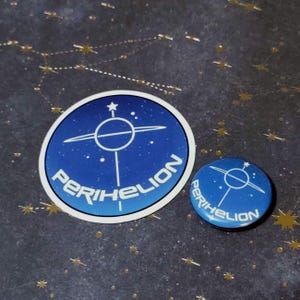 May include: A blue and white sticker and button featuring the word "Perihelion" and a celestial design. The sticker is round, and the button is a smaller, circular version of the sticker design. The background is dark blue with gold star constellations.