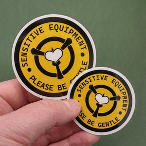 以下が含まれることがあります： 「SENSITIVE EQUIPMENT PLEASE BE GENTLE」の文字が入った2つの丸いステッカー。ステッカーは黄色で、黒と白のアクセントがあります。デザインには白いハートと黒いプロペラのような形が含まれています。