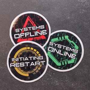 以下が含まれることがあります： 技術をテーマにしたデザインの丸いステッカー3枚。「SYSTEMS OFFLINE」と白い文字と赤い三角形で書かれたステッカーが1枚。「SYSTEMS ONLINE」と緑色で書かれたステッカーが1枚。3枚目のステッカーには「INITIATING RESTART」と黄色で書かれています。