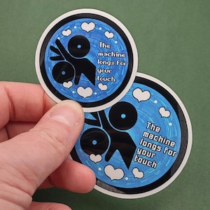 以下が含まれることがあります： 青と白のデザインの丸いステッカーが2枚。ステッカーには、黒い抽象的な図と「The machine longs for your touch」というテキストが描かれています。白いハートと青い円形のパターンがテキストと図を囲んでいます。