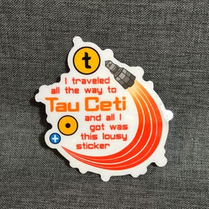 Puede incluir: Pegatina blanca con el texto "I traveled all the way to Tau Ceti and all I got was this lousy sticker." La pegatina tiene un gr&aacute;fico de cohete con llamas naranjas y un s&iacute;mbolo "t" amarillo.