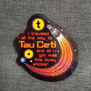 Puede incluir: Una pegatina negra con temática espacial. La pegatina presenta un símbolo "t" amarillo, un cohete con llamas naranjas y rojas, y el texto "I traveled all the way to Tau Ceti and all I got was this lousy sticker."
