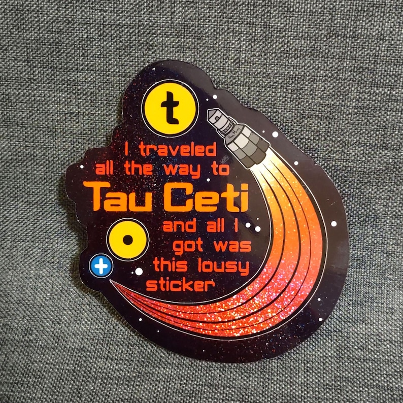 Puede incluir: Una pegatina negra con tem&aacute;tica espacial. La pegatina presenta un s&iacute;mbolo "t" amarillo, un cohete con llamas naranjas y rojas, y el texto "I traveled all the way to Tau Ceti and all I got was this lousy sticker."