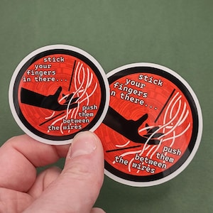 以下が含まれることがあります： 赤い背景に白い文字で「stick your fingers in there... push them between the wires」と書かれた2つの丸いステッカー。中央には黒い手のグラフィックがあります。ステッカーは緑色の背景にあります。
