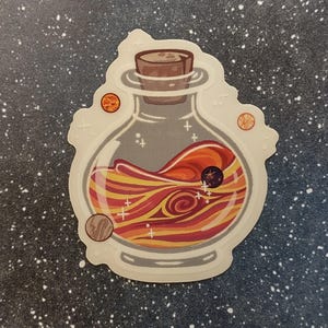 May include: A sticker featuring a glass bottle with a cork stopper, filled with swirling orange, red, and yellow liquid. Small planets and stars surround the bottle, creating a cosmic theme. The sticker has a white border.