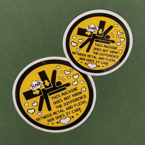 以下が含まれることがあります： 黄色い背景と黒い縁取りの丸いステッカー2枚。ステッカーには、機械に押しつぶされる人のグラフィックと「THIS MACHINE DOES NOT KNOW THE DIFFERENCE BETWEEN METAL AND FLESH, NOR DOES IT CARE.」というテキストが表示されています。