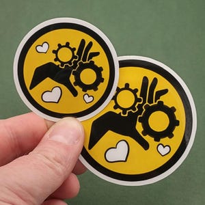 以下が含まれることがあります： 黄色い背景、黒い縁取り、2つの歯車を持つ手の黒いグラフィックが特徴の2つの丸いステッカー。白いハートが中央の画像に散らばっています。ステッカーは手で持たれています。