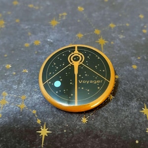 Puede incluir: Un pin redondo con borde naranja y centro negro. Presenta un diseño dorado y la palabra "Voyager". El diseño incluye un mapa estelar estilizado con un punto turquesa. El pin está sobre un fondo oscuro con patrones de estrellas doradas.
