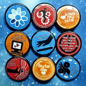 May include: A collection of nine circular pin badges with diverse designs and text. The badges display a variety of colours, including blue, orange, and red. Some badges feature text such as "Kiss my bits" and "Byte me". Other badges have illustrations of a flower, a computer, and a person.