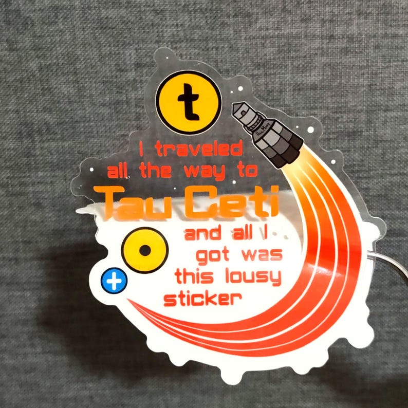 Puede incluir: Una pegatina transparente con un dise&ntilde;o de cohete y el texto "I traveled all the way to Tau Ceti and all I got was this lousy sticker." La pegatina tiene llamas naranjas y blancas, un c&iacute;rculo amarillo y un signo m&aacute;s.