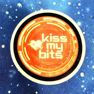 以下が含まれることがあります： オレンジと黄色の円形デザインの中に、白で「kiss my bits」と書かれた丸いステッカー。 黒い縁と白い縁があり、白い点のある青い背景に配置されています。