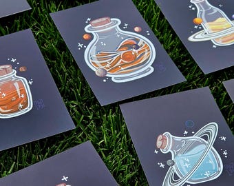 May include: A collection of rectangular art prints on a dark background, each featuring a glass bottle with a cork stopper. The bottles contain swirling liquids in various colors, surrounded by small planetary spheres, rings, and white cross-shaped stars.