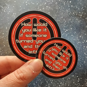以下が含まれることがあります： 「How would you like it if someone turned you and then left?」という白文字が赤と黒のデザインで描かれた丸いステッカー2枚。ステッカーは手で持たれています。