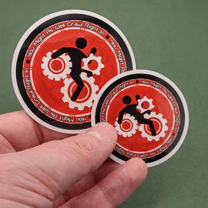 以下が含まれることがあります： 赤と黒のデザインの丸いステッカーが2枚。白い歯車と相互作用する黒い棒人間が特徴で、「You Might As Well Crawl Right In」というテキストで囲まれています。ステッカーは手で持たれています。