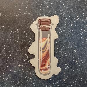 May include: A sticker featuring a glass vial with a cork stopper. The vial contains a swirling mixture of orange, yellow, and purple colors. The sticker has a white border with small cross-shaped designs.