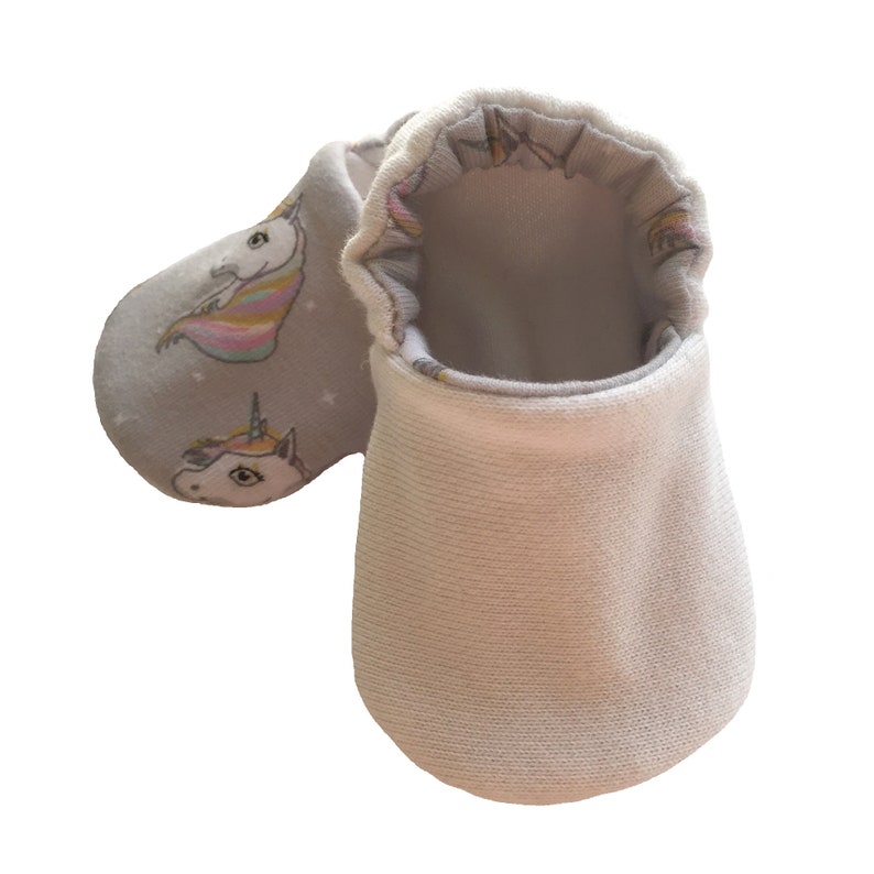 May include: A pair of gray baby booties with a white unicorn print. The booties are made of soft, comfortable fabric and have a white cuff.
