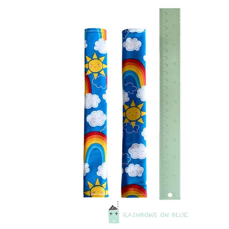 May include: Two blue fabric seatbelt covers with a repeating pattern of smiling suns, rainbows, and fluffy white clouds. A green ruler is next to the covers. The text "Rainbows on Blue" is at the bottom.