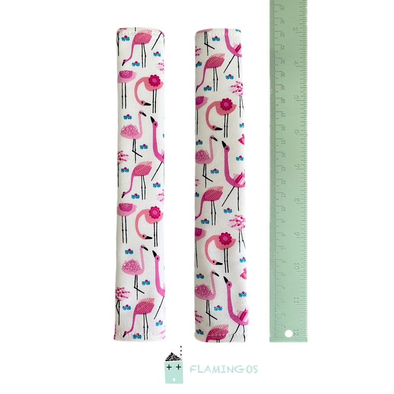 May include: Two pink flamingo patterned fabric sleeves, each measuring 10 inches long, are shown next to a ruler. The sleeves are designed to protect furniture from scratches and wear.