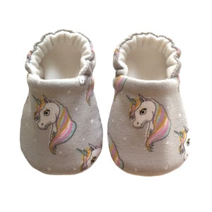 May include: A pair of gray baby booties with a unicorn print. The booties have a white fleece lining and a white elastic band around the top.