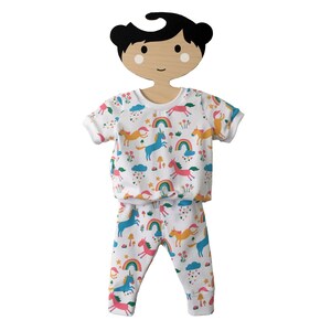 May include: White baby pajamas with a unicorn, rainbow, and cloud print. The pajamas are on a wooden doll hanger.
