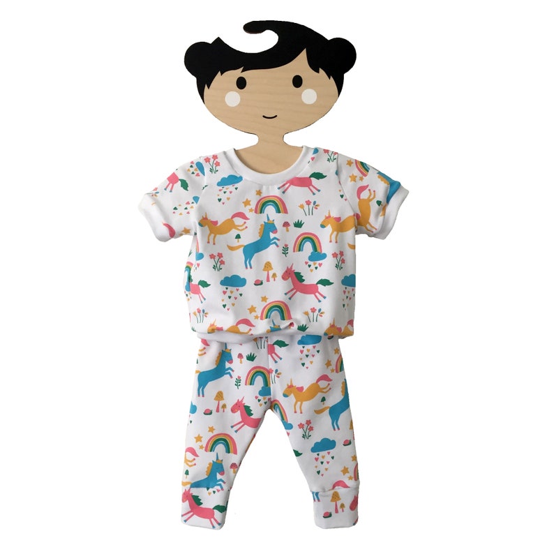 May include: White baby pajamas with a unicorn, rainbow, and cloud print. The pajamas are on a wooden doll hanger.