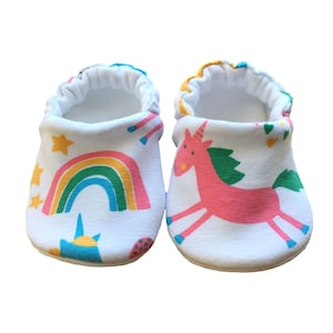 May include: A pair of white baby booties with a colorful unicorn and rainbow design. The booties have elastic at the top for a snug fit.