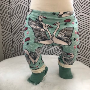 Op de afbeelding: Groene baby leggings met een wit zwanenpatroon. De zwanen zijn gestileerd en hebben een geometrisch ontwerp. De leggings hebben een witte boord aan de enkel.