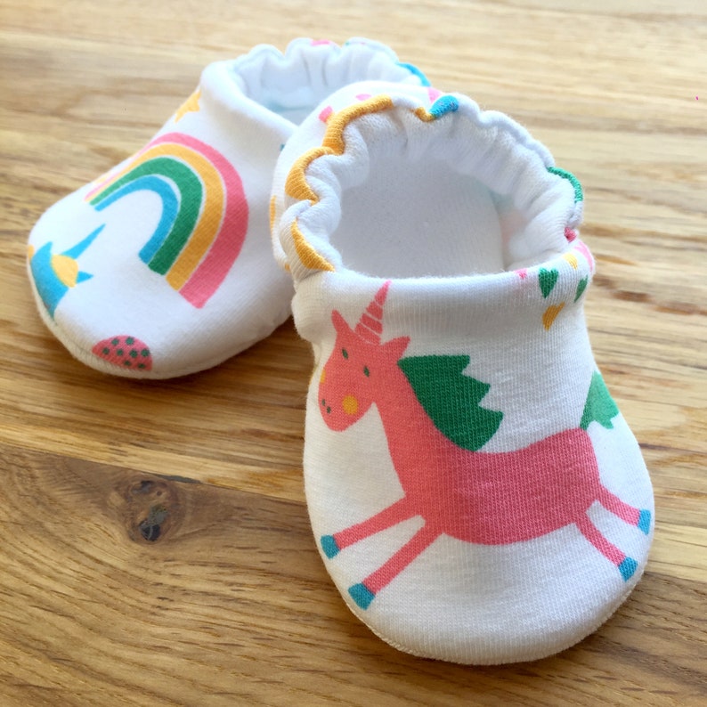 May include: A pair of white baby booties with a rainbow and unicorn design. The booties are made of soft cotton and have a comfortable fit.