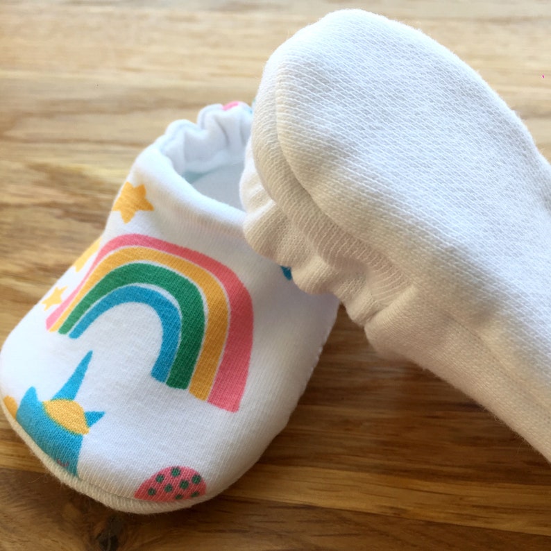 May include: A pair of white baby booties with a rainbow, stars, and a unicorn design. The booties are made of soft cotton and have a fold-over cuff.