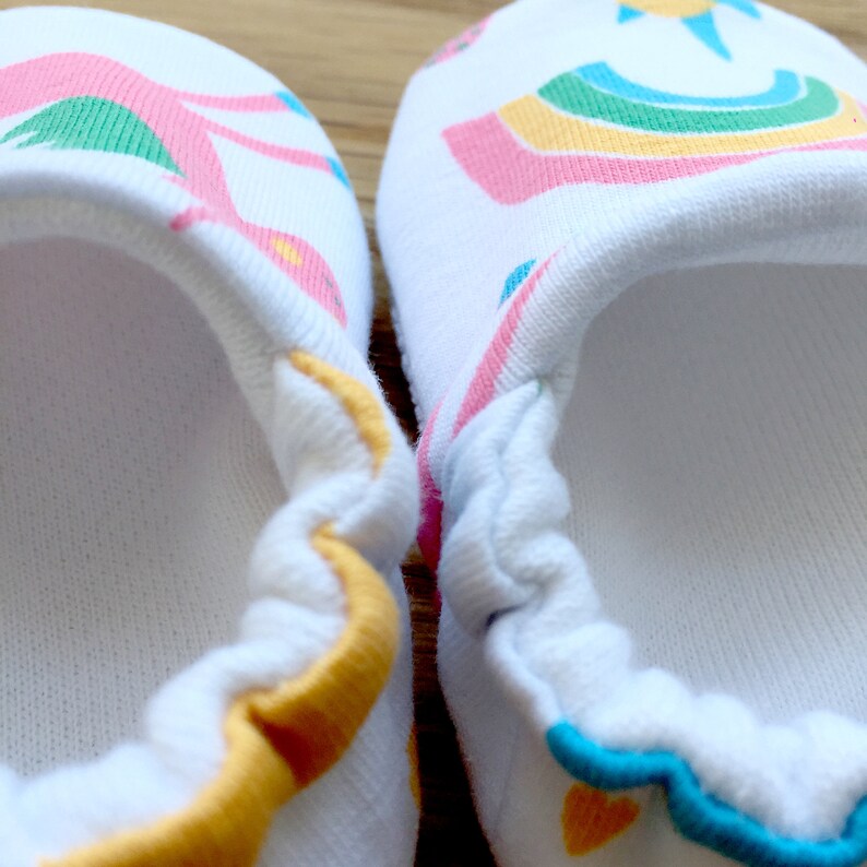 May include: A pair of white baby booties with a colorful rainbow and sun design. The booties have a yellow, orange, and blue trim.