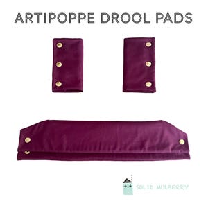May include: Three-piece set of Artipoppe drool pads in a deep purple hue. The set includes two smaller pads with three gold-colored buttons each, and a larger, curved pad with two buttons on each end. The text "ARTIPOPPE DROOL PADS" is at the top.