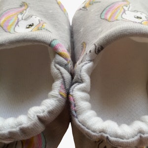 May include: A pair of gray baby booties with a white unicorn print. The booties have a white elastic band around the opening.