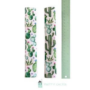 May include: Two white fabric drawer pulls with a cactus print. The fabric is white with green and blue watercolor cactus illustrations. The pulls are approximately 11 inches long.