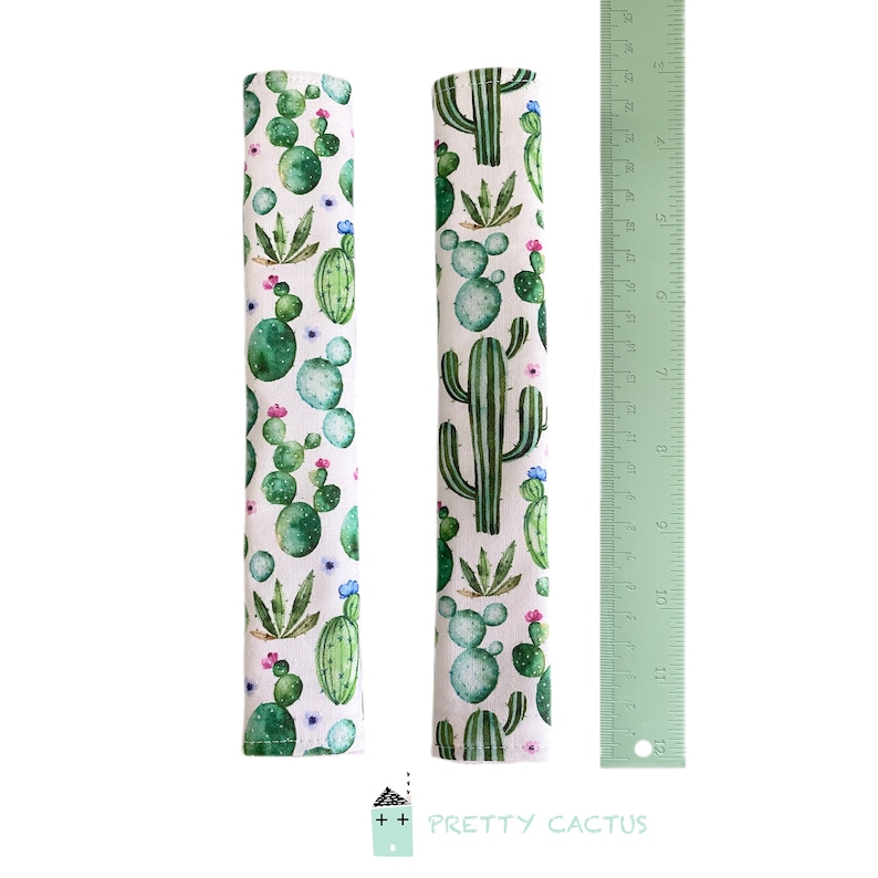 May include: Two white fabric drawer pulls with a cactus print. The fabric is white with green and blue watercolor cactus illustrations. The pulls are approximately 11 inches long.