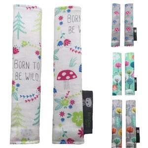May include: Set of two white fabric car seat belt covers with a woodland theme. One cover features a green tree and the text "Born to be Wild". The other cover features a pink mushroom, green leaves, and flowers. Both covers have a small black label with a bear illustration and the text "Bella Cash Designs".