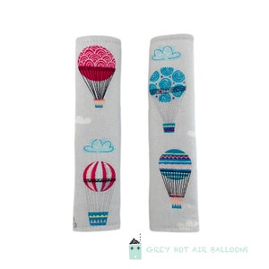 May include: Two gray fabric car seat belt covers with a hot air balloon pattern. The pattern features blue, pink, and white hot air balloons with clouds in the background. The text "GREY HOT AIR BALLOONS" is printed on the bottom of the image.