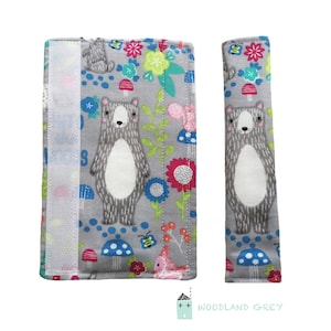 May include: A pair of gray fabric car seat belt strap covers with a woodland animal print featuring bears, mushrooms, flowers, and bees. The strap covers have a white velcro closure.