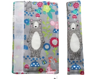 May include: A pair of gray fabric car seat belt strap covers with a woodland animal print featuring bears, mushrooms, flowers, and bees. The strap covers have a white velcro closure.