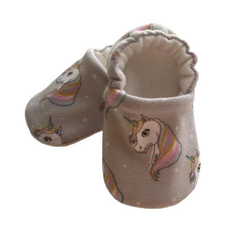 May include: A pair of gray baby booties with a unicorn print. The unicorns have rainbow manes and tails. The booties have a white lining.