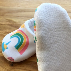 May include: A pair of white baby booties with a rainbow, sun, and star design. The booties are made of soft cotton and have a white sole.