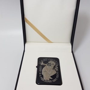 Op de afbeelding: Zwarte aansteker met een zilveren gravure van een vrouw die een plant vasthoudt en de tekst "Mary Green - The Herb is a Precious Gift from God". De aansteker zit in een zwarte doos met een witte binnenkant.